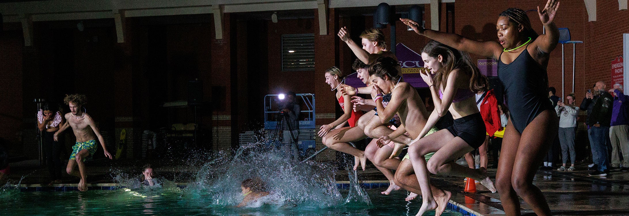 A group of students jumping into a swimming pool at night, making splashes