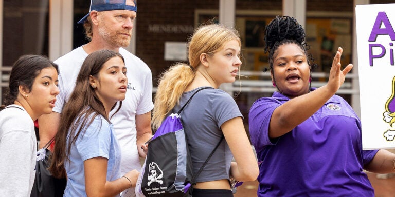 A Pirate Navigator in a purple collared shirt holding an ‘Ask a Pirate’ sign helps direct prospective students around campus.