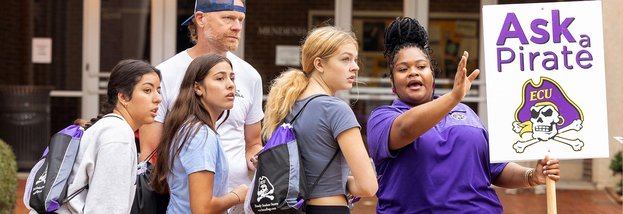 A Pirate Navigator in a purple collared shirt holding an ‘Ask a Pirate’ sign helps direct prospective students around campus.