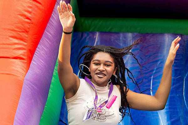 A student in a tank top and sweat pants slides down an inflatable slide at a campus event in late summer