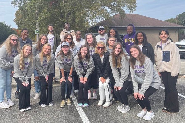 A group of nearly 20 students wearing E C U sweatshirts pose for a photo in a parking lot.